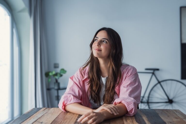 Adult woman sitting at a table looking thoughtfully toward a window representing the experience of undiagnosed ADHD in women