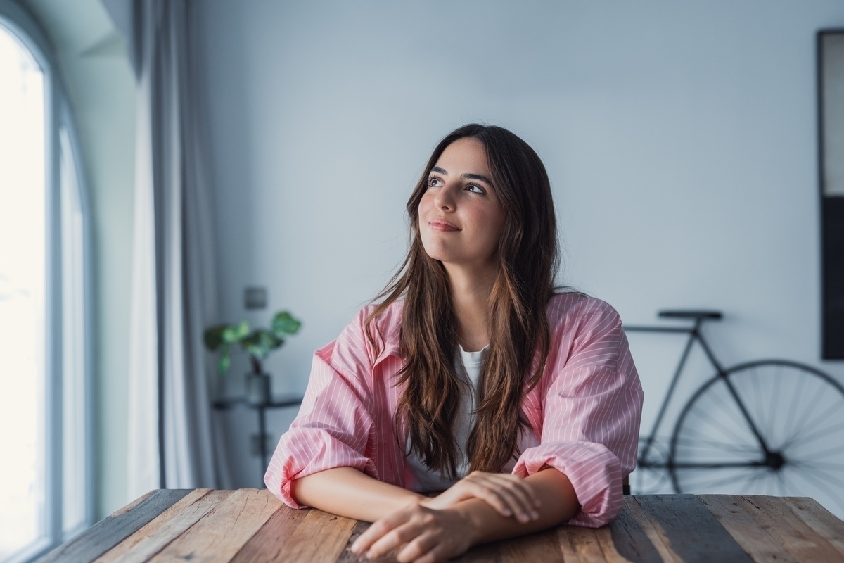 Adult woman sitting at a table looking thoughtfully toward a window representing the experience of undiagnosed ADHD in women