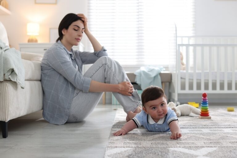 Exhausted mother sitting on the floor beside her baby representing the emotional experience of postpartum depression