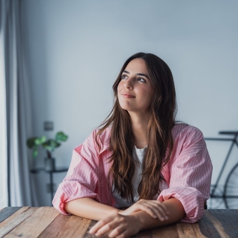 Adult woman sitting at a table looking thoughtfully toward a window representing the experience of undiagnosed ADHD in women