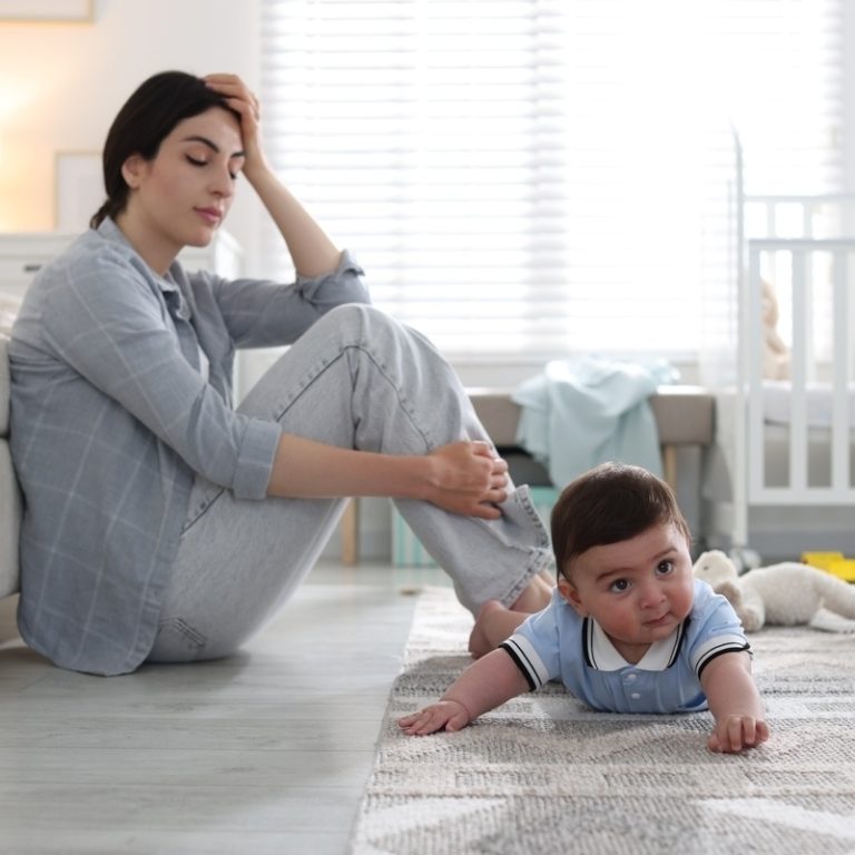 Exhausted mother sitting on the floor beside her baby representing the emotional experience of postpartum depression